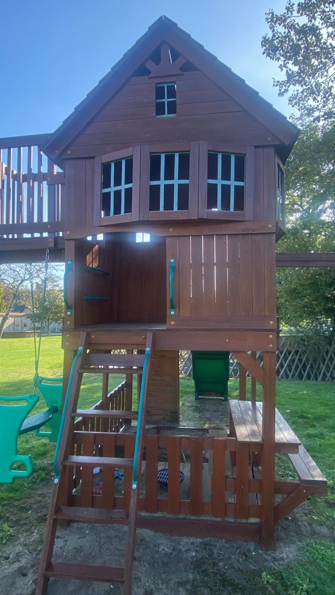 Structure de jeu en bois avec toboggan et étagère, située dans un jardin verdoyant.