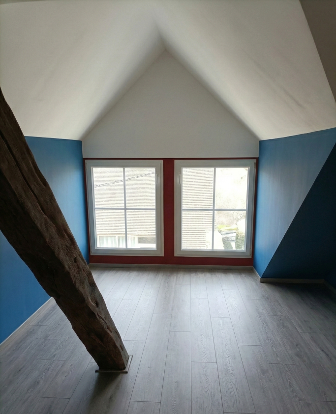 Pièce avec murs bleu et blanc, fenêtres rouges, sol en bois et poutre apparente.